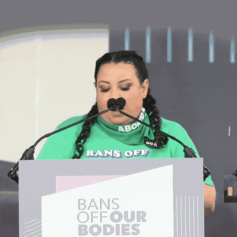 Video gif. Woman wearing a bright green t-shirt speaks emphatically at a podium above a sign that reads, "Bans off our bodies." She says, "Today is day one of an uprising to protect abortion rights, day one of our feminist future, day one of a summer of rage."