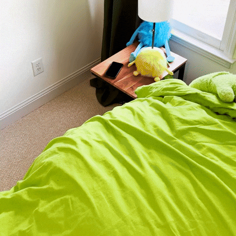 Video gif. Person in a giant smiley lime green mascot costume pulls back the lime green covers of their bed and stretches their arms out then side to side. 