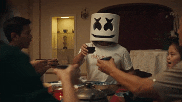 Celebrity gif. Marshmello, the DJ, sits with a family and they all have cups up as they're about to clink and cheers.