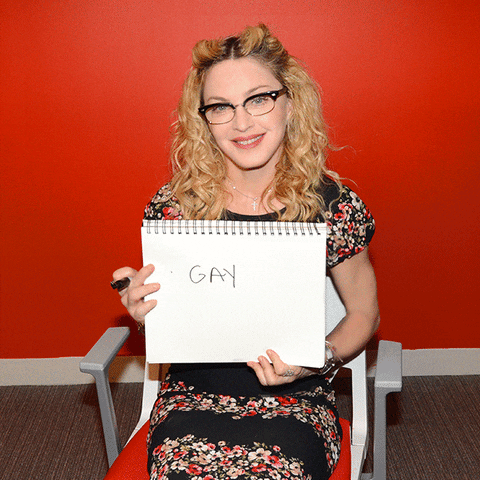 Photo gif. Madonna sits in a chair smiling and holding up a notebook on which the word "gay" is written.