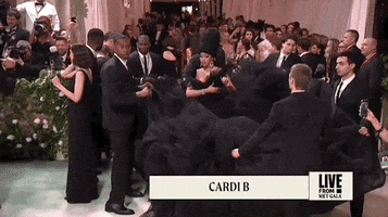 Met Gala 2024 gif. Cardi B walks down in her Windowsin strapless black gown assisted by six attendants holding up her giant trumpet skirt so she can walk down the red carpet. She's wearing a black matching beehive-style headpiece. She's gesturing out to the attendants like she's trying to coordinate all of their movements together.