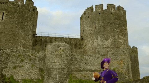 Gif of a princely looking white man in a purple outfit complete with a feather in his hat trotting along on a hobby horse in front of a castle.
