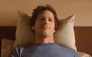 Movie gif. Andy Samberg as Nyles in Palm Springs lays in bed and makes a cranking gesture with his fingers near the sides of his face, turning his straight face into a sarcastic, toothy smile.