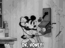 Disney gif. Original black-and-white cartoon style Mickey Mouse stands on the back of a chair speaking into an old-fashioned telephone on the wall, blowing a kiss and saying, "Ok, honey!" which appears as text.