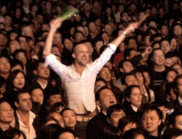 Video gif. A man standing up in a crowd dances as he waves glow sticks in the air. 