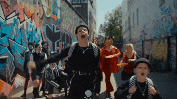 Music video gif. In a clip from the music video for "Little Brother" by Yungblud, Dominic Richard Harrison, dressed in mostly black with a checkered backpack, does a cheerful march down an alley covered in graffiti as kids in Halloween costumes follow behind and a person in a nun costume rides by on a skateboard.