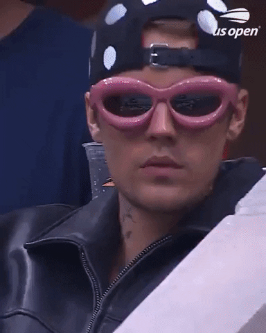 Video gif. Justin Bieber wears a leather jacket, a polka dot hat backwards baseball hat, and hot pink thick rimmed sunglasses as he watches a game from the stands.