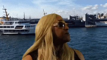 Celebrity gif. Robert E Blackmon wears sunglasses and a straight blonde wig, giving a wide smile for a camera as it pans across boats in the water. A circle springs around his face and text appears, "Special Guest Star: Thot."
