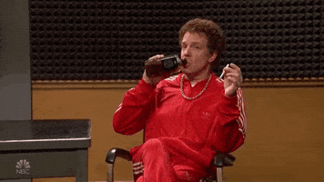 SNL gif. In front of a wall with studio "egg crate" soundproofing, Jason Sudeikis sits in an office chair wearing a red tracksuit and a necklace. He points at us while drinking straight from a liquor bottle.