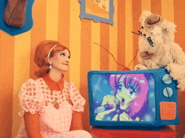 Video gif. Woman in a 50s style wig sits in front of a small vintage TV that has an anime girl on it singing into a microphone. She gives a knowing smile up at a white furry puppet perched on top of the TV who is leaning down to switch the TV off. Text, "Sweet dreams."
