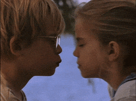 Movie gif. Anna Chlumsky as Vada and Macaulay Caulkin as Thomas in My Girl are in front of a lake and share a small smooch before separating with surprise on their faces.