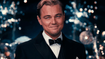 Movie gif. Leonardo DiCaprio as Jay Gatsby in The Great Gatsby raises a champagne glass in a celebratory toast as fireworks explode behind him.