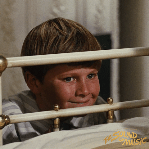Movie gif. Duane Chase as Kurt Von Trapp in The Sound of Music raises up from behind the railing of a bed, looking over it with a slow, goofy grin.