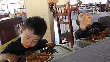 Video gif. Two young toddlers sit in high chairs with plates of spaghetti in front of them. Red Sauce is all over their faces. One kid bobs his head around and tries to keep his eyes open like he’s fighting sleep. The Other has his head all the way back and is shoveling noodles into his mouth.