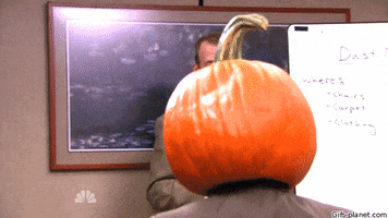 The Office gif. Leading a staff meeting in a conference room, Paul Lieberstein as Toby calls on Rainn Wilson as Dwight, who's raising his hand and wearing a jack-o-lantern on his head.