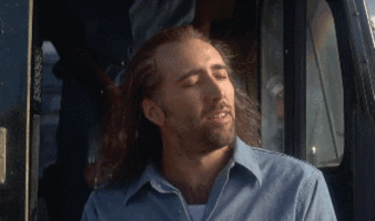 Digital compilation gif. Nicholas Cage as Cameron in the movie, "Con Air" in a scene where he's exiting the prison van, chained to the inmate behind him. He feels the sun on his face and the wind in his hair, really getting lost and feeling the moment, giving off a somewhat unsettling sense of euphoria. The scene changes to a highly edited animated scene featuring just his head bouncing around in a fast moving galaxy filled with stars and supernovas flashing all around him. 