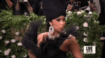 Met Gala 2024 gif. Closeup of the top of Cardi B's black strapless Windowsin gown decorated with a black wing motif. She holds up her long pointy emerald green nails to blow a kiss and touches the end of her black fabric and tulle wings that extend up the front of the bodice to just above her shoulders.