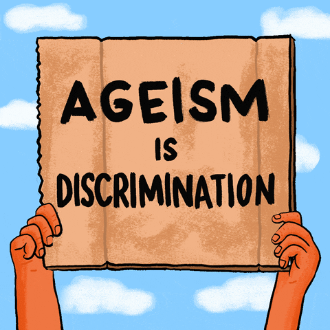 Illustrated gif. Hands hold up a cardboard sign straight up in the air like they are taking part in a protest. The sign has handwritten text in bold black letters that reads, "Ageism is discrimination."
