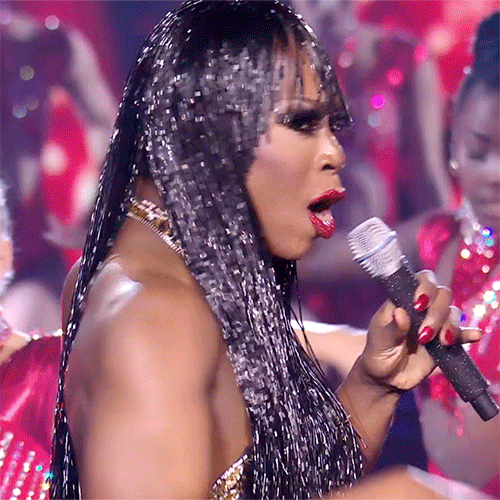 Reality TV gif. Aria B Cassadine on Queen of the Universe, performing in a sparkly headdress with microphone in hand, points at us as she sings, "You!"