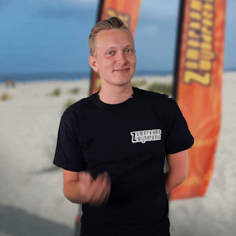 Video gif. A man standing on a beach has one hand behind his back and the other gestures for us to continue talking, trying to find the point in what we're saying.