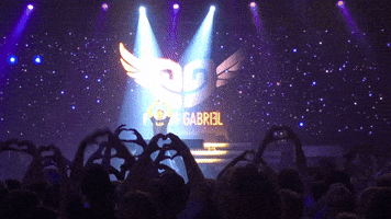 Video gif. Music artist Pieter Gabriel stands on stage in front of a cosmic backdrop with a twirling winged, heart-shaped logo. He interacts with an adoring crowd of fans, who are making hearts with their hands and recording him.