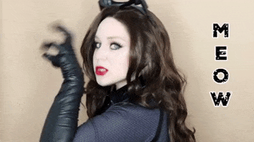 Celebrity gif. Lillee Jean dressed as Catwoman, faces us in profile, as she makes a claw with her hand and scratches down, staring at us with her teeth showing. Text, "Meow."