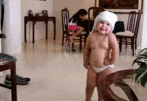 Video gif. A toddler wearing only a diaper and a towel on their head dances proudly, wiggling their body and shaking their hips.