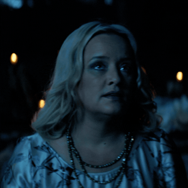 TV gif. Kiernan Sipka as Sabrina in Chilling Adventures of Sabrina looks up at the big glowing full moon, and then we see, from above, a group of people lying on the ground around a fire, in a circle, reaching to link hands.