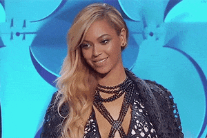 Celebrity gif. Beyonce is on stage smiling and she hears something that makes her fold her lips in and look down, suddenly getting flirtatious.