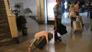 Video gif. In the lobby of a busy hotel, a man rolls his luggage with a little girl dead asleep laying on top of it. He just looks at her and then keeps moving, wondering if she’ll ever wake up.