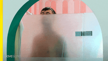 Reality TV gif. Man on Love Island USA stands in the shower and peeks over the glass door.