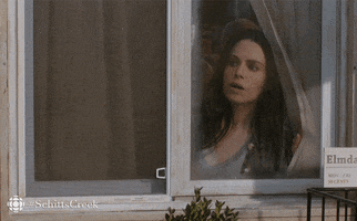 Schitt's Creek. Emily Hampshire as Stevie holds a curtain aside and gazes out a window with interest, waiting.