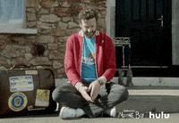 TV gif. Chris O'Dowd as Seán in Moone Boy sits on a front step beside an old suitcase and looks down at his watch in exasperation.