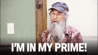 Video gif. A man with a long white beard and vintage aviator glasses hold his palm to his chest while saying, "I'm in my prime."