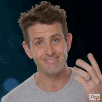 Reality TV gif. Joey McIntyre from Rock This Boat New Kids on the Block, smiles a toothy grin at us and touches his fingers to his dimples.