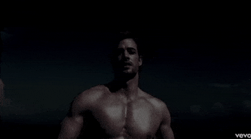 Music Video gif. From Jennifer Lopez's video for "I'm Into You," William Levy walks toward us in slow motion, shirtless on a beach at night.