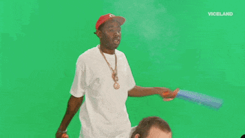 Celebrity gif. Tyler the Creator stands in front of a greenscreen and swings a flimsy toy sword to fan away smoke, which makes text reading, "Welcome Back" appear.