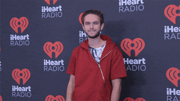 Celebrity gif. DJ Zedd points finger guns at us, walking the red carpet in front of the I Heart Radio step-and-repeat.