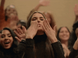 Music video gif. A woman in a crowd of others uses both hands to send twice as many air kisses.