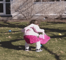 Video gif. Young girl holds a pink bucket and picks up an easter egg. She tosses it up without much thought and turns around while it’s in the air. Somehow, the egg still makes it into the basket.