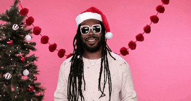 Celebrity gif. Rapper Dram wears big square sunglasses and a santa hat as he stands in front of a Christmas tree. He spreads his arms out and smiles as he says, “Merry Christmas, ho ho ho!”