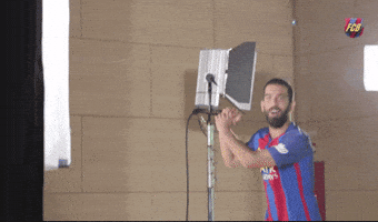 Sports gif. Arda Turan in a FC Barcelona jersey claps his hands and smiles as he stands on a photoshoot set.