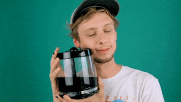 Video gif. A man holds up and leans his face against a coffee pitcher, smiling slightly with his eyes closed.