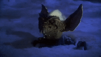 Movie gif. Character Stripe from Gremlins makes an evil face before sinking down and disappearing into the snow. 