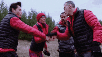 Reality TV gif. Standing outdoors in puffy jackets, four contestants on American Grit put their hands together in a gesture of teamwork and say, “Go team!”