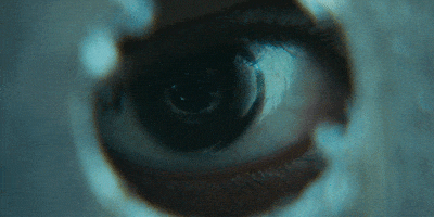 Movie gif. Close-up of a person's eye peering through a small round opening from the horror movie "It Comes at Night."