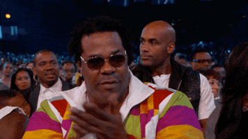 Celebrity gif. Busta Rhymes sits in the audience at the BET awards. He wears tinted sunglasses and he claps and nods along with the rest of the crowd. 