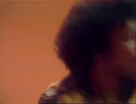 TV gif. In a scene from Soul Train, a smiling man with a moustache, big curly hair, and a cardigan tied around his shoulders spreads his arms in a cheerful dance against a solid orange background.