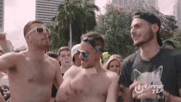 Reality TV gif. Group of shirtless men wearing sunglasses jumping and dancing among a larger crowd at a concert.