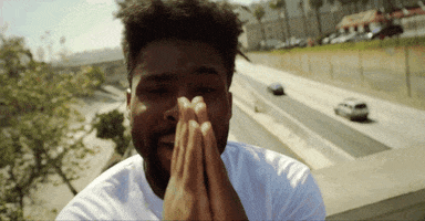 Video gif. Man stands on a bridge over a highway. He holds his hands up in front of his face in a prayer position, and bobs his head side to side.